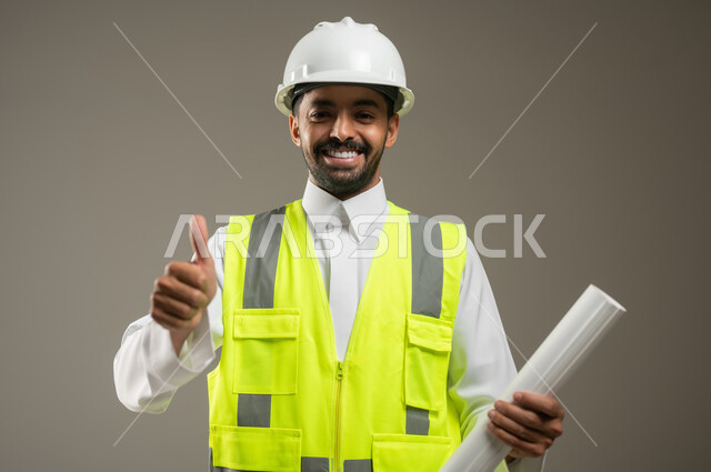 The concept of architectural construction and engineering, the quality of art in design and construction technology, a close-up portrait of a smiling Saudi Arabian Gulf engineer wearing a helmet and protection vest, pointing with a thumb up with the quality sign, holding building and construction plans in his hand, gray background, sensitive content