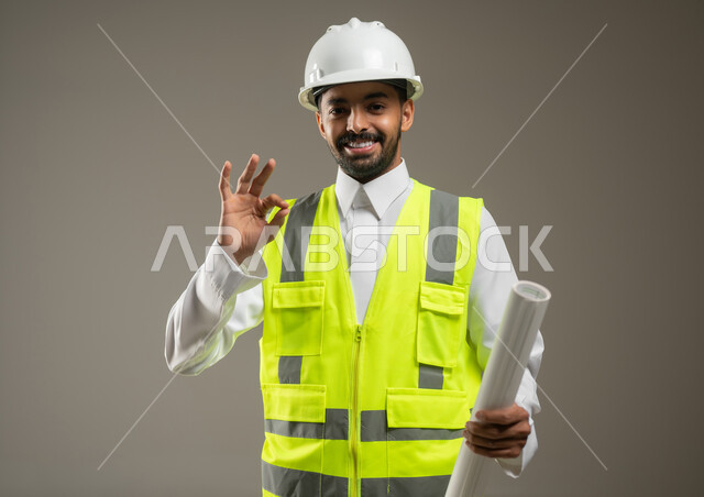مفهوم الانشاءات المعمارية والهندسة، جودة الفن في تقنية التصميم والبناء، بورتريه مقرب لمهندس عربي خليجي سعودي مبتسم يرتدي الخوذه و سترة الحماية يشير بيده للأعلى بعلامه الجوده، يحمل بيده مخططات البناء والتشييد، خلفية رمادية، محتوى حساس