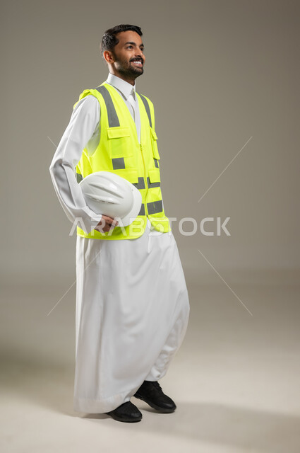 بورتريه لكامل الجسم لمهندس عربي خليجي سعودي مبتسم، يرتدي ستره الحمايه يحمل بيده خوذه بيضاء، يقوم بالتحرك والمشي، ينظر الى شئ ما بايماءات التفاؤل، التنقل والذهاب من مكان ما، مفهوم الذهاب والعوده، خلفيه رماديه، استخدام حساس، محتوى حساس