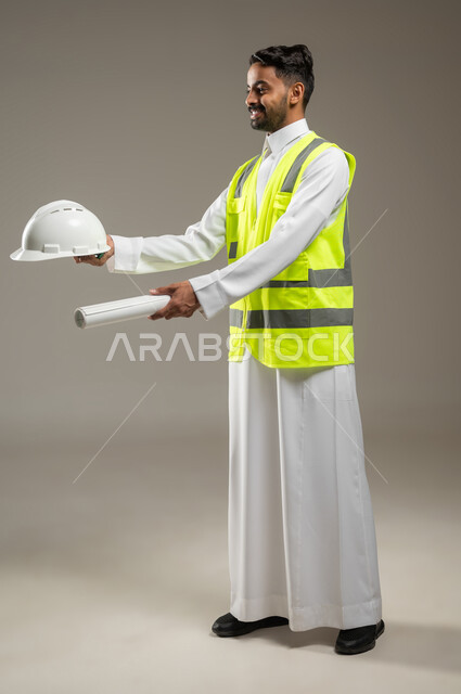 معلومات عن مشاريع هندسية، أوراق خاصة بالعمل، بورتريه بالطول الكامل لرجل خليجي عربي سعودي يرتدي الثوب التقليدي مع سترة الحماية الخاصة بالعمل يحمل بيديه خوذه الحمايه ومخطط البناء، يشير بهما الى مكان ما، خلفية رمادية , استخدام حساس , محتوى حساس