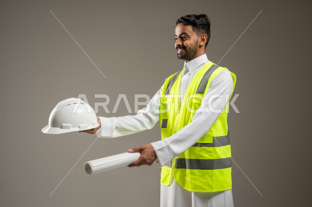 معلومات عن مشاريع هندسية، أوراق خاصة بالعمل، بورتريه مقرب لرجل خليجي عربي سعودي يرتدي الثوب التقليدي مع سترة الحماية الخاصة بالعمل يحمل بيديه خوذه الحمايه ومخطط البناء، يشير بهما الى مكان ما، خلفية رمادية , استخدام حساس , محتوى حساس