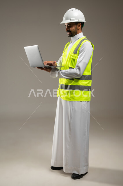 بورتريه جانبي بالطول الكامل لمهندس عربي خليجي سعودي يحمل جهاز اللابتوب المحمول ، مهندس انشاءات يرتدي بدلة السلامة في العمل ، خوذة العمل ، يعمل على جهازه المحمول، مشروع بناء و مقاولات، العمل عن بعد، خلفيه رماديه، استخدام حساس، محتوى حساس