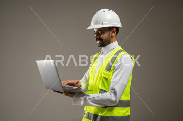 Close-up portrait of a Saudi Gulf Arab engineer holding a laptop, a construction engineer wearing a safety suit at work, a work helmet, working on his mobile device, a construction and contracting project, remote work, gray background, sensitive use, sensitive content