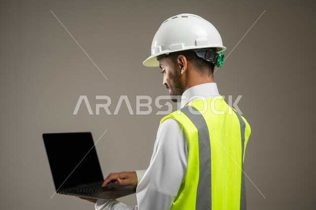 A close-up portrait of a Saudi Gulf Arab engineer holding a laptop, a construction engineer wearing a safety suit at work, a work helmet, a construction and contracting project, remote work, gray background, sensitive use, sensitive content.