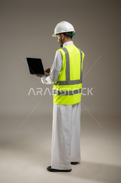 بورتريه جانبي خلفي بالطول الكامل لمهندس عربي خليجي سعودي يحمل جهاز اللابتوب المحمول ، مهندس انشاءات يرتدي بدلة السلامة في العمل ، خوذة العمل ، مشروع بناء و مقاولات، العمل عن بعد، خلفيه رماديه، استخدام حساس، محتوى حساس