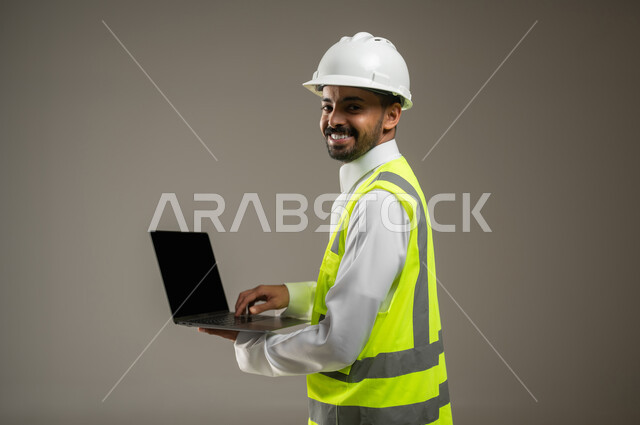 Close side portrait of a Saudi Arabian Gulf engineer holding a laptop, a construction engineer wearing a safety suit at work, a work helmet, looking at the camera with a smile, a construction and contracting project, remote work, gray background, sensitive use, sensitive content