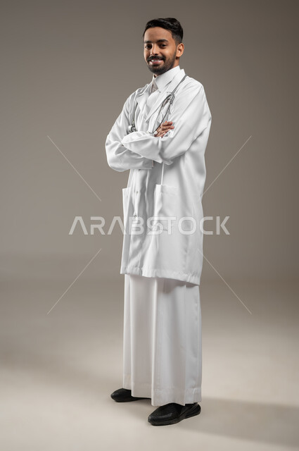 A Saudi Gulf Arab doctor standing sideways with his hands folded, full-length portrait of a health practitioner wearing a white medical coat, placing his stethoscope around his neck, looking at the camera with a smile, gray background, sensitive content, sensitive use