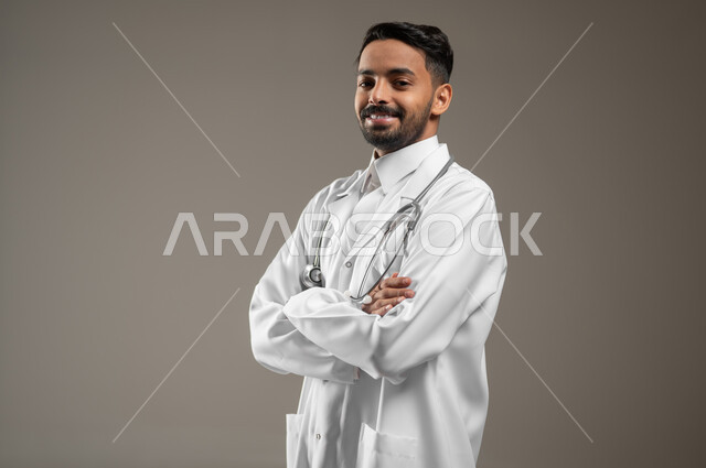 A Saudi Gulf Arab doctor standing sideways with his hands folded, close-up portrait of a health practitioner wearing a white medical coat and wearing his stethoscope around his neck looking at the camera with a smile, gray background, sensitive content, sensitive use