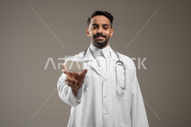 الوقوف والاشارة إلى بطاقة عمل تعريفية شخصية، بورتريه مقرب لطبيب عربي خليجي سعودي مبتسم يرتدي الثوب السعودي والمعطف الطبي الابيض ، يمسك بطاقة بيضاء فارغة, موك اب ابيض، خلفية رمادية، محتوى حساس، استخدام حساس