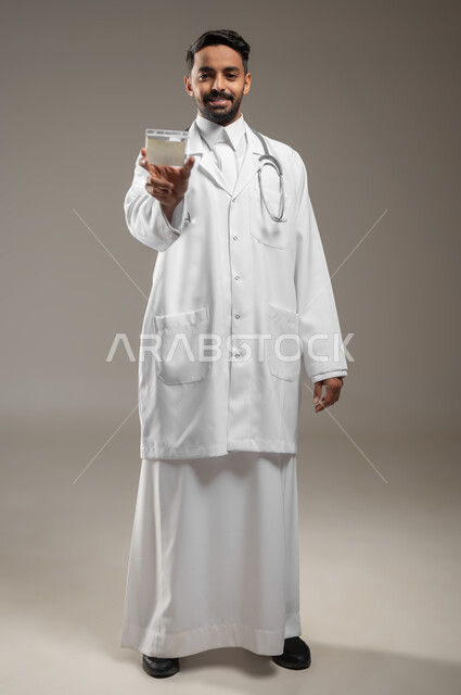 الوقوف والاشارة إلى بطاقة عمل تعريفية شخصية، بورتريه لطبيب عربي خليجي سعودي مبتسم يرتدي الثوب السعودي والمعطف الطبي الابيض ، يمسك بطاقة بيضاء فارغة, موك اب ابيض صورة الجسم بالطول الكامل, خلفية رمادية، محتوى حساس، استخدام حساس