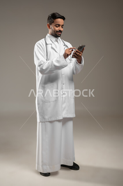 تقديم الخدمات للمرضى بكل كفاءه, اجراء الاستشارات الطبيه عن بعد, استخدام الجوال في البحث والتطوير، بورتريه بالطول الكامل لطبيب عربي خليجي سعودي يرتدي المعطف الابيض يضع سماعته حول رقبته، يمسك بجهازه المحمول، خلفيه رماديه، محتوى حساس، استخدام حساس