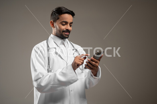 تقديم الخدمات للمرضى بكل كفاءه, اجراء الاستشارات الطبيه عن بعد, استخدام  الجوال في البحث والتطوير، بورتريه مقرب لطبيب عربي خليجي سعودي يرتدي المعطف الابيض يضع سماعته حول رقبته، يمسك بجهازه المحمول، خلفيه رماديه، محتوى حساس، استخدام حساس