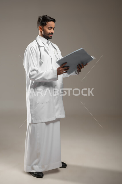 قراءة التحاليل والتقارير الطبية، صورة الجسم بالكامل لطبيب عربي خليجي سعودي يرتدي المعطف الأبيض يضع السماعات الطبيه حول رقبته، يحمل بيده أوراق وملفات المرضى، العمل في القطاع الصحي في المملكة العربية السعودية، تقديم الرعاية الصحية للمرضى، خلفيه رماديه، محتوى حساس