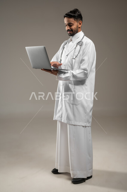 طبيب عربي خليجي سعودي يرتدي المعطف الابيض الطبي يضع سماعته الطبيه حول رقبته، بورتريه جانبي بالطول الكامل لممارس صحي يمسك بيده جهازا محمولا ويقوم بالعمل عليه، العمل عن بعد، تقنيه الحاسوب، خلفيه رماديه، محتوى حساس