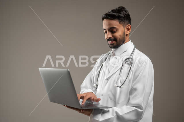 طبيب عربي خليجي سعودي يرتدي المعطف الابيض الطبي يضع سماعته الطبيه حول رقبته، بورتريه مقرب جانبي لممارس صحي يمسك بيده جهازا محمولا ويقوم بالعمل عليه، العمل عن بعد، تقنيه الحاسوب، خلفيه رماديه، محتوى حساس