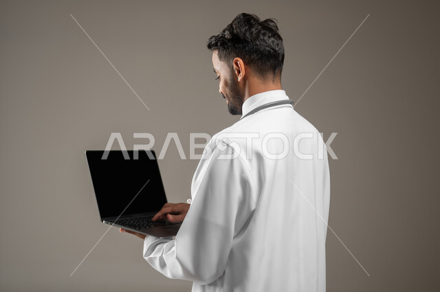 طبيب عربي خليجي سعودي يرتدي المعطف الابيض الطبي ويضع سماعته الطبيه حول رقبته، بورتريه مقرب جانبي خلفي لممارس صحي يمسك بيده جهازا محمولا ويقوم بالعمل عليه، العمل عن بعد، تقنيه الحاسوب، خلفيه رماديه، محتوى حساس