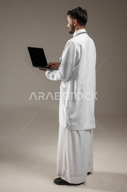 طبيب عربي خليجي سعودي يرتدي المعطف الابيض الطبي ويضع سماعته الطبيه حول رقبته، بورتريه جانبي خلفي بالطول الكامل لممارس صحي يمسك بيده جهازا محمولا ويقوم بالعمل عليه، العمل عن بعد، تقنيه الحاسوب، خلفيه رماديه، محتوى حساس