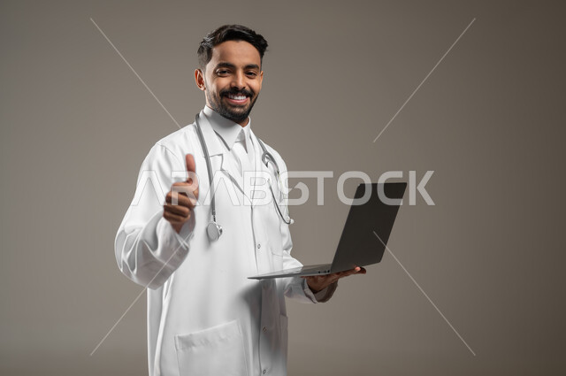 طبيب عربي خليجي سعودي يمسك بيده جهازا محمولا يشير بإصبعه الإبهام لأعلى بإيماءات وجه تدل على القبول والموافقة والإعجاب، بورتريه مقرب لممارس صحي يرتدي المعطف الابيض الطبي ويضع سماعته الطبيه ينظر الى الكاميرا بابتسامه، خلفيه رماديه، محتوى حساس