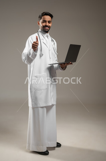 a Saudi Arabian Gulf doctor holding a portable device in his hand, pointing with his thumb upward with facial gestures indicating acceptance, approval and admiration, a full-body photo of a health practitioner wearing a white medical coat and wearing his stethoscope, looking at the camera with a smile, gray background, sensitive content