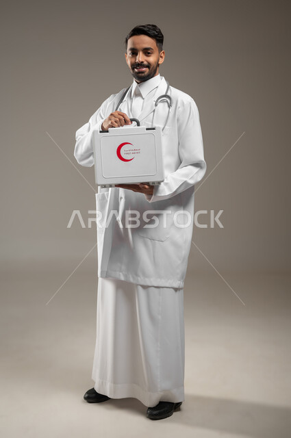 التعامل مع الإصابات الشائعة وحالات الطوارئ، تقديم الخدمات الصحية الطارئة للمرضى، بورتريه لطبيب عربي سعودي خليجي يرتدي الثوب التقليدي و المعطف الطبي يحمل بيده حقيبة الاسعافات الأولية، الأدوات والمعدات الطبية اللازمة، صورة الجسم الكامل، خلفية رماديه، محتوى حساس