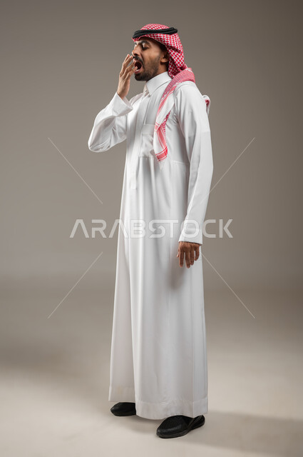 النعاس والحاجه الى النوم، بورتريه لرجل عربي سعودي خليجي يرتدي الزي السعودي، يضع يده على فمه بإيماءات وجه ويد تدل على النعاس , الإحساس بالتعب والتثاؤب , الحاجة للنوم والراحة , الكسل والخمول، صوره الجسم بالطول الجانبي الكامل، خلفيه رماديه، استخدام حساس