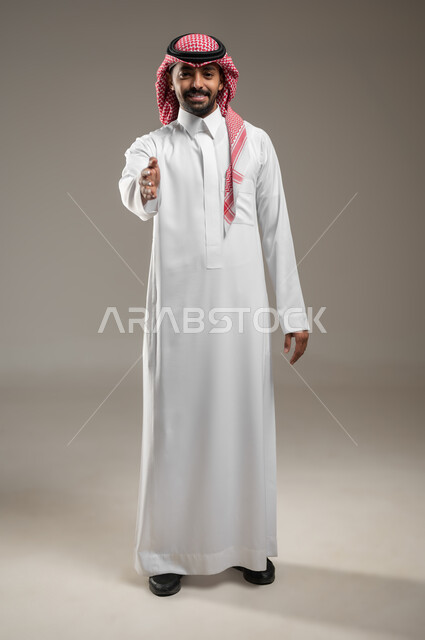 إيماءات السعادة والتحية والسلام والترحيب، بورتريه لرجل عربي خليجي سعودي يرتدي الشماغ السعودي والثوب الابيض يمد يده بإشارة المصافحة بايماءات الترحيب والسعاده، رجل سعودي مبتسم، صوره الجسم بالطول الكامل، حركة يد وإشارات، خلفيه رماديه، محتوى حساس