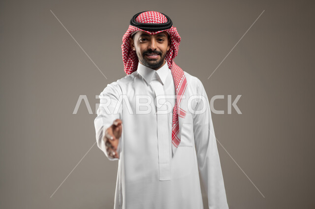 إيماءات السعادة والتحية والسلام والترحيب، بورتريه مقرب لرجل عربي خليجي سعودي يرتدي الشماغ السعودي والثوب الابيض يمد يده بإشارة المصافحة بايماءات الترحيب والسعاده، رجل سعودي مبتسم، حركة يد وإشارات، خلفيه رماديه، محتوى حساس