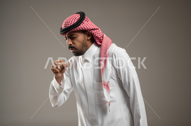 Symptoms of the Corona virus (Covid-19), asking for help to go to the doctor to develop a treatment plan, contracting a cold, close-up portrait of a young Arab Gulf Saudi man wearing the Saudi uniform with gestures indicating coughing and pain, feeling sick, tired and exhausted, gray background, sensitive content