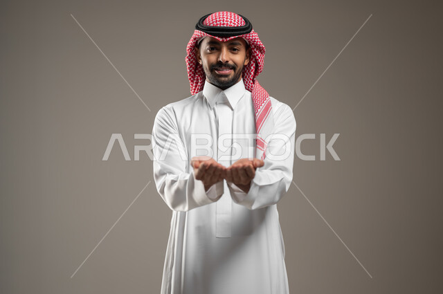 إمساك وإحتواء عنصر هولوجرام، تقنية الهولوجرام ثلاثية الابعاد، بورتريه مقرب لرجل عربي خليجي سعودي يرتدي الزي السعودي، يفتح يديه لعرض عنصر هولوجرامي, ايماءات يدين ووجه تدل على السعادة, خلفية رماديه، استخدام حساس، محتوى حساس