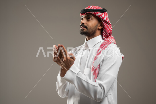 اشارات وجه ويدين تدل على الدعاء، التضرع الى الله, بورتريه لرجل عربي خليجي سعودي ينظر الى الاعلى، الحمد والشكر و طلب العون, الرغبه في تحقيق الأمنيات, يرتدي الزي السعودي يرفع يديه للاعلى، يطلب العون والمغفره من الله، خلفيه رماديه، استخدام حساس، محتوى حساس