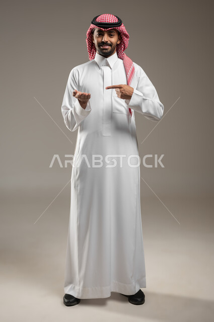 النظر إلى اليدين بإيماءات السعادة والسرو، التوجه و الإشارة لشيء ما، مفهوم لغة الجسد، بورتريه لرجل عربي خليجي سعودي مبتسم يرتدي الزي السعودي يشير باصبعه السبابة إلى يده الأخرى، لغه الاشارات، صورة لكامل الجسم، خلفية رمادية، محتوى حساس، استخدام حساس