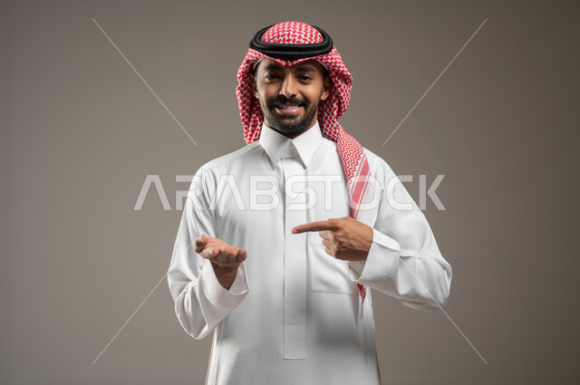 النظر إلى اليدين بإيماءات السعادة والسرو، التوجه و الإشارة لشيء ما، مفهوم لغة الجسد، بورتريه مقرب لرجل عربي خليجي سعودي مبتسم يرتدي الزي السعودي يشير باصبعه السبابة إلى يده الأخرى، لغه الاشارات، خلفية رمادية، محتوى حساس، استخدام حساس
