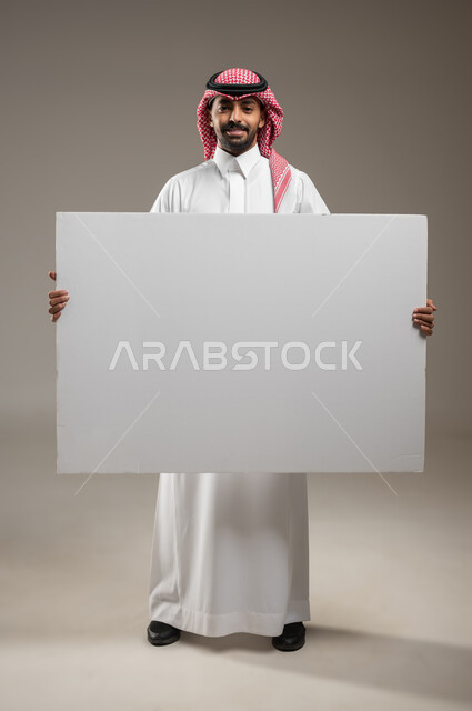 استخدام لوحات توضيحية تعريفية، إعلانات وعروض تسويقية، موك اب مربع أبيض كبير، بورتريه لرجل عربي خليجي سعودي يرتدي الزي السعودي يحمل بيده لوحه بيضاء مربعه فارغة ينظر الى الكاميرا، صوره الجسم بالطول الكامل، خلفية رماديه، محتوى حساس، استخدام حساس