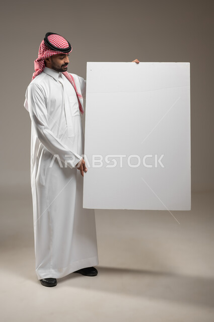 استخدام لوحات توضيحية تعريفية، إعلانات وعروض تسويقية، موك اب مربع أبيض كبير، بورتريه لرجل عربي خليجي سعودي يرتدي الزي السعودي يحمل بيده لوحه بيضاء مربعه فارغة ينظر إليها وهو واقفا بالطول الجانبي، صوره الجسم بالطول الكامل، خلفية رماديه، محتوى حساس، استخدام حساس