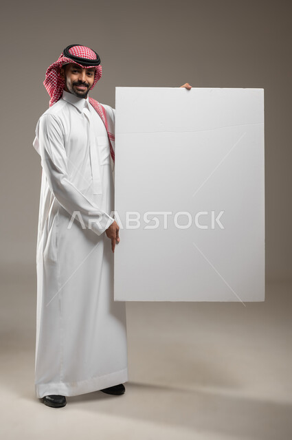 استخدام لوحات توضيحية تعريفية، إعلانات وعروض تسويقية، موك اب مربع أبيض كبير، بورتريه لرجل عربي خليجي سعودي يرتدي الزي السعودي يحمل بيده لوحه بيضاء مربعه فارغة ينظر إلى الكاميرا بالطول الجانبي بايماءات السعاده، صورة الجسم بالكامل، خلفية رماديه، محتوى حساس