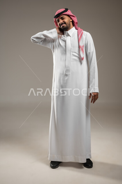 رجل عربي سعودي خليجي يرتدي الزي السعودي، يضع يده على رقبته بايماءات وجه و يد تدل على التعب والارهاق، آلام الرقبه والعمود الفقري ، شد عضلي، التعب والارهاق, الحاجه الى العلاج, صوره الجسم بالطول الكامل، خلفيه رماديه، استخدام حساس، محتوى حساس