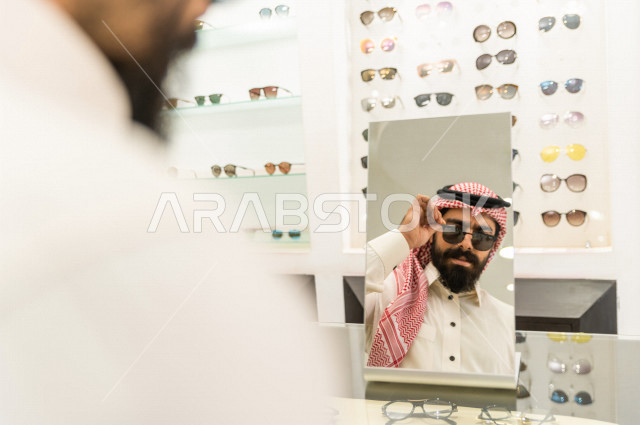 شاب عربي سعودي يقف امام المرآة , ويجرب نظارة شمسية تلائم ملامح وجهه في محل نظارات .  