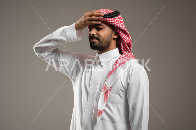 الشعور بالتعب والارهاق، بورتريه مقرب لرجل عربي سعودي خليجي يرتدي الزي السعودي يضع يده على رأسه ، إيماءات وجه ويد تدل على الشعور بالصداع، ألم بالرأس، التعب والإرهاق ، مشاكل جسدية ، حالة مرضية، خلفيه رماديه، محتوى حساس، استخدام حساس