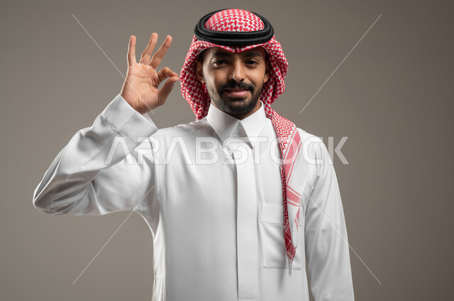السرور والسعادة بإنجاز المهام، بورتريه مقرب لشاب عربي خليجي سعودي مبتسم يرتدي الزي السعودي ينظر إلى الكاميرا، رفع اليدين بعلامة الجودة والامتياز، الدقة في تحقيق أهداف العمل, خلفيه رماديه، محتوى حساس، استخدام حساس