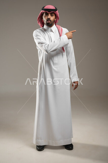 الاشارة نحو شيء وتحديد هدف ما، بورتريه  لرجل عربي خليجي سعودي يرتدي الشماغ والثوب السعودي يشير بأصبع السبابة على مكان معين بإيماءات الصدمة والذهول، التعجب والاستغراب والاندهاش ، صوره الجسم بالطول الكامل، خلفية رماديه، استخدام حساس، محتوى حساس