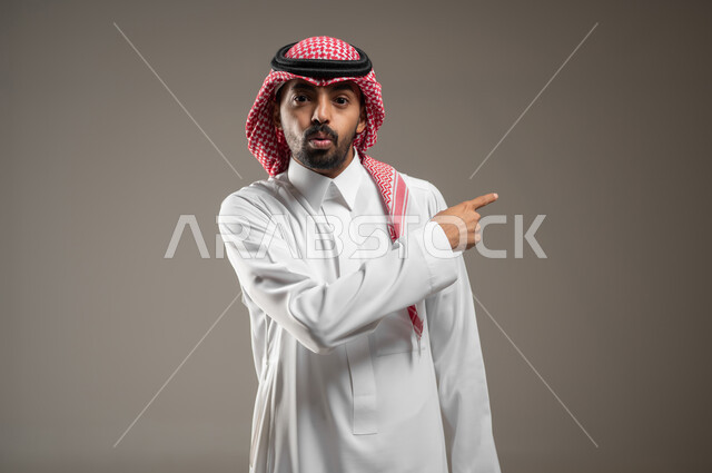 الاشارة نحو شيء وتحديد هدف ما، بورتريه مقرب لرجل عربي خليجي سعودي يرتدي الشماغ والثوب السعودي يشير بأصبع السبابة على مكان معين بإيماءات الصدمة والذهول، التعجب والاستغراب والاندهاش ، خلفية رماديه، استخدام حساس، محتوى حساس