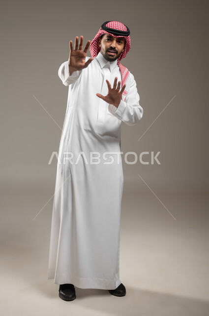 Prohibition from doing something, portrait of a Saudi Gulf Arab young man wearing Saudi uniform, pointing forward with his hands, angry facial gestures and hands indicating prohibition and stopping, gestures and movements, reaction and signals, full-length body image, gray background, sensitive use, sensitive content