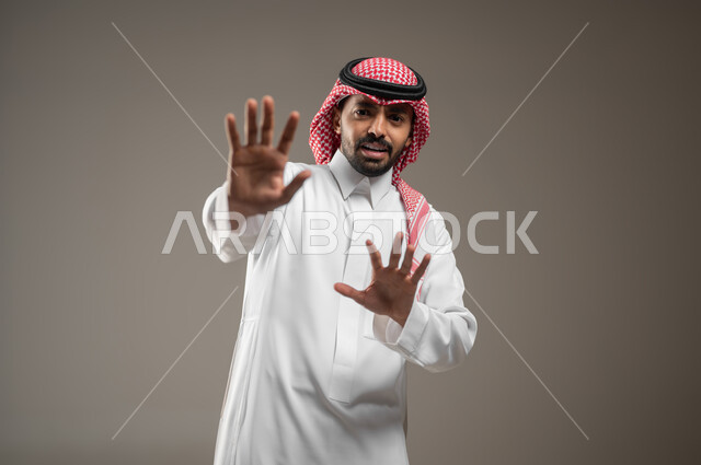 النهي عن فعل شئ ما، بورتريه مقرب لشاب عربي خليجي سعودي يرتدي الزي السعودي، يشير بيديه الى الامام، إيماءات وجه غاضبة ويدين تدل على المنع والتوقف، ايماءات وحركات، رد فعل واشارات، خلفيه رماديه، استخدام حساس، محتوى حساس