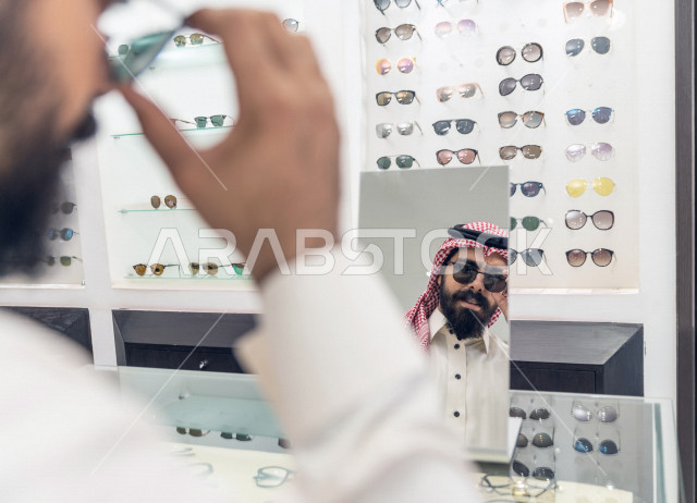 شاب عربي سعودي يقف امام المرآة , ويجرب نظارة شمسية تلائم ملامح وجهه في محل نظارات .  