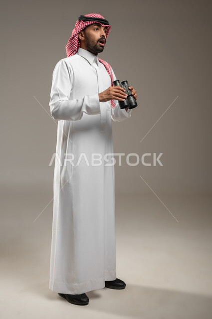 بورتريه لرجل عربي خليجي سعودي يرتدي الزي السعودي، يحمل بيده المنظار ثنائي العينين ( التلسكوب ) ، بإيماءات وتعابير وجه مختلفه، الاستكشاف والمغامرة ، التعجب و الاستغراب، صوره الجسم بالطول الكامل، خلفيه رماديه، استخدام حساس، محتوى حساس