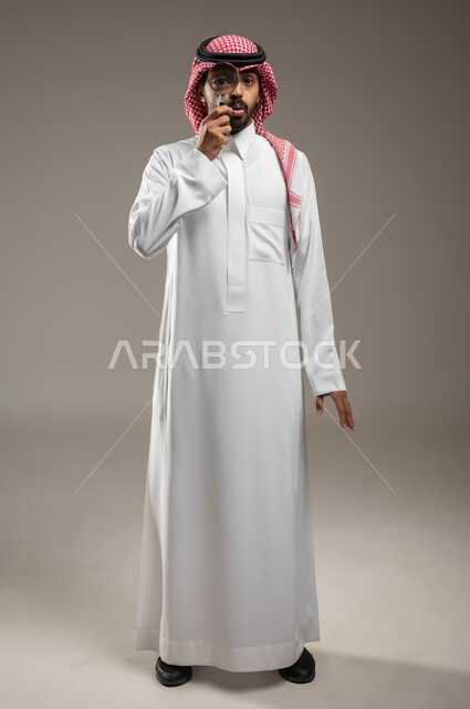 Portrait of a Saudi Gulf Arab man wearing Saudi uniform, looking through a magnifying glass, medical optical tools, close-up view using a magnifying glass, full-length body portrait, gray background, sensitive content, sensitive use