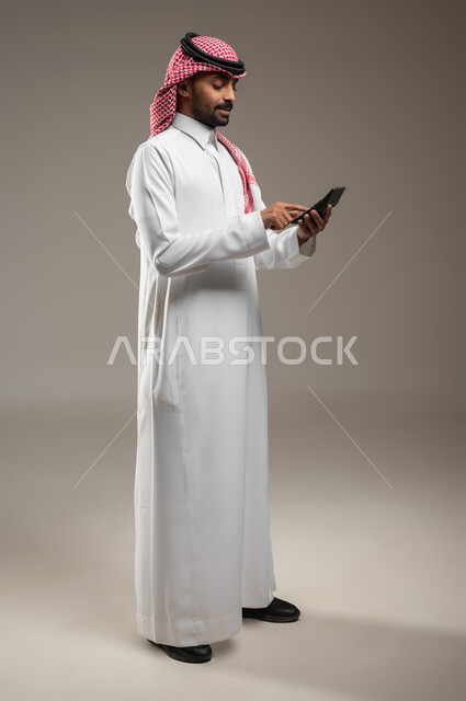 بورتريه لرجل عربي خليجي سعودي يرتدي الزي السعودي، يحمل بيده آلة حاسبة ، يشير باصبعه الى ازرار الاله الحسابيه، استخدام الآلة الحاسبة في إجراء العمليات الحسابية ، مفهوم أدوات مكتبية ، صوره الجسم بالطولل الكامل، خلفية رماديه، استخدام حساس، محتوى حساس
