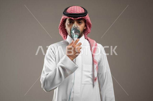 Providing home health services to the patient, treating cases of shortness of breath, ensuring comfort for the respiratory muscles using a respirator, a close-up portrait of a Saudi Arabian Gulf man wearing a Saudi uniform inhaling shortness of breath medications using an oxygen mask, gray background, sensitive content