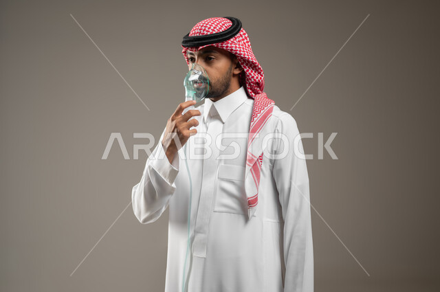 تقديم الخدمات الصحية المنزلية للمريض، معالجة حالات ضيق التنفس، تأمين الراحة للعضلات التنفسية باستخدام جهاز التنفس الصناعي، بورتريه مقرب لرجل عربي سعودي خليجي يرتدي الزي السعودي يقوم باستنشاق أدوية ضيق التنفس بواسطه كمامه الاكسجين، خلفيه رماديه، محتوى حساس