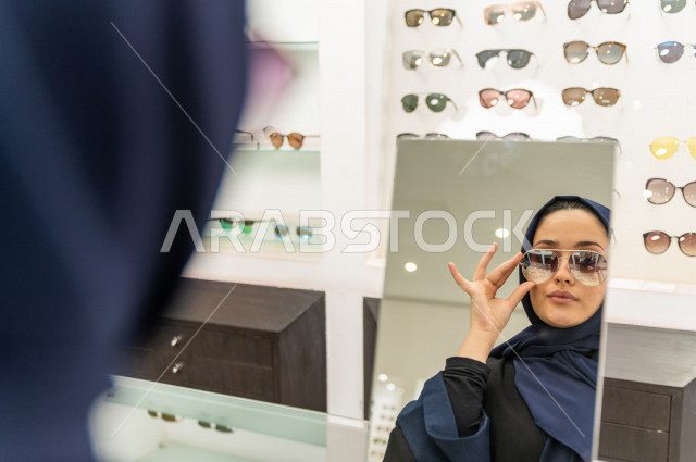 امرأة خليجية تقف امام المرآه  لاختيار نظارة شمسية مناسبة في محل نظارات , اختيار عصري للنظارات الشمسية